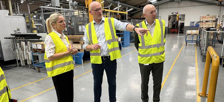 Tre personer i gule sikkerhedsveste står i en produktionshal og taler sammen under en rundvisning.